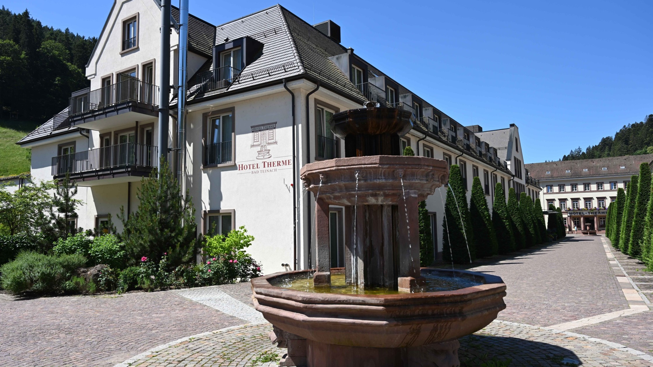 Hotelaußenansicht mit Springbrunnen, gepflegtem Weg und grünen Bäumen im Sommer