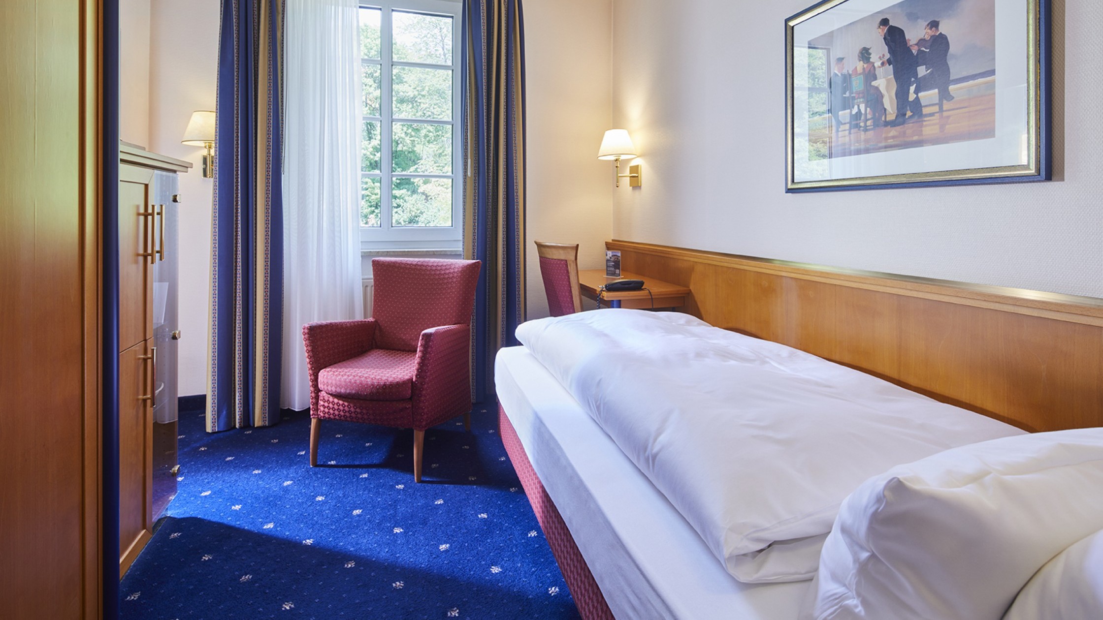 Gemütliches Einzelzimmer mit blauem Teppich, rotem Sessel und Schreibtisch am Fenster