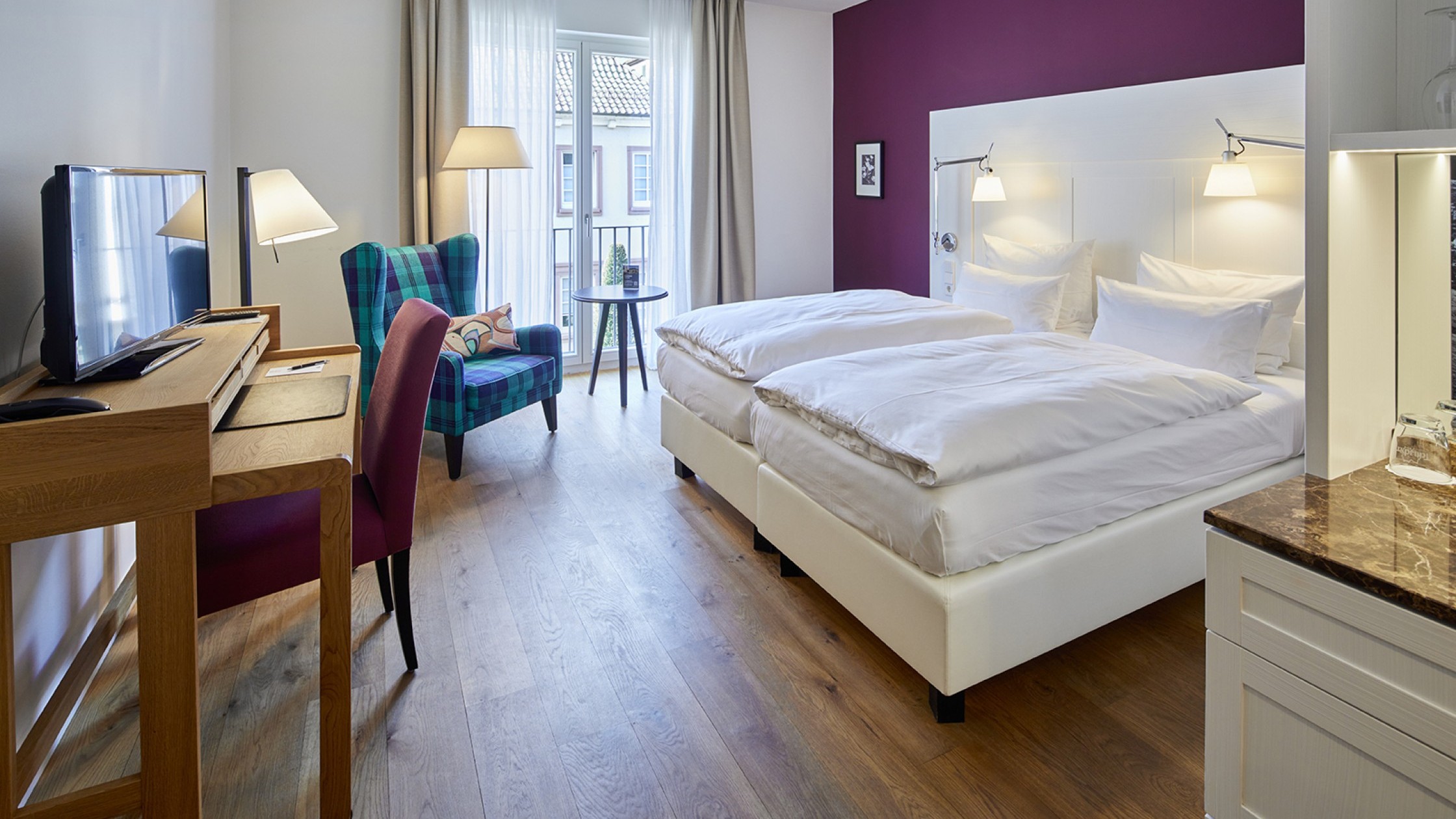 Modernes Doppelzimmer mit violetter Akzentwand, hellem Holzboden und Sitzbereich am Fenster